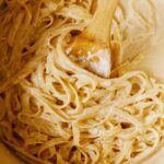 A big pot full of freshly cooked fettuccine alfredo with a wooden spoon in it.