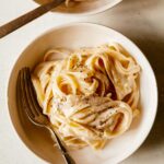 Two bowls of fettuccine alfredo with forks and fresh cracked pepper on top.