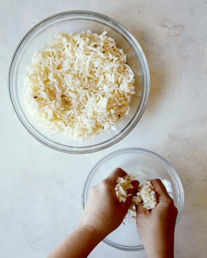 Thawed hash browns in a mixing bowl with hands squeezing out the moisture.