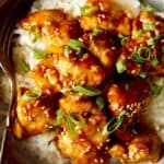 A bowl of sesame chicken with sesame seeds and green onions over rice with a fork.