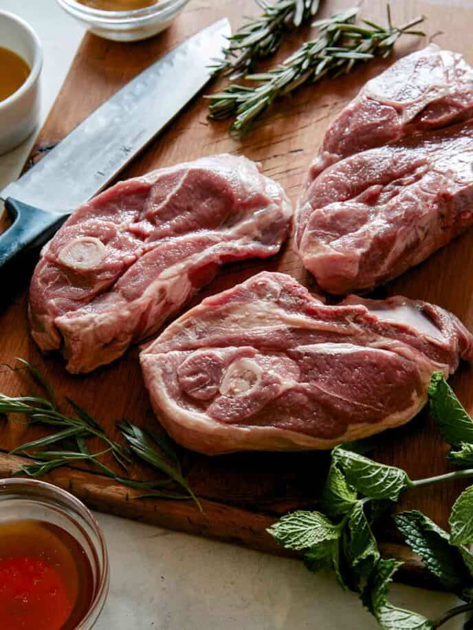three lamb shoulders on a cutting board with a knife and some herbs.