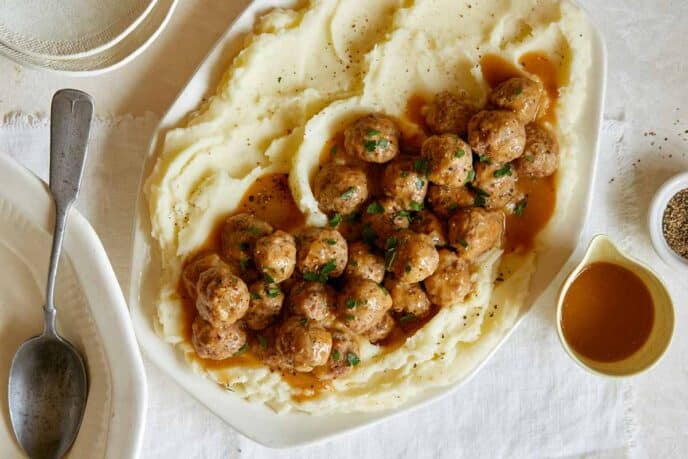 A platter of Swedish meatballs over mashed potatoes with plates to serve.