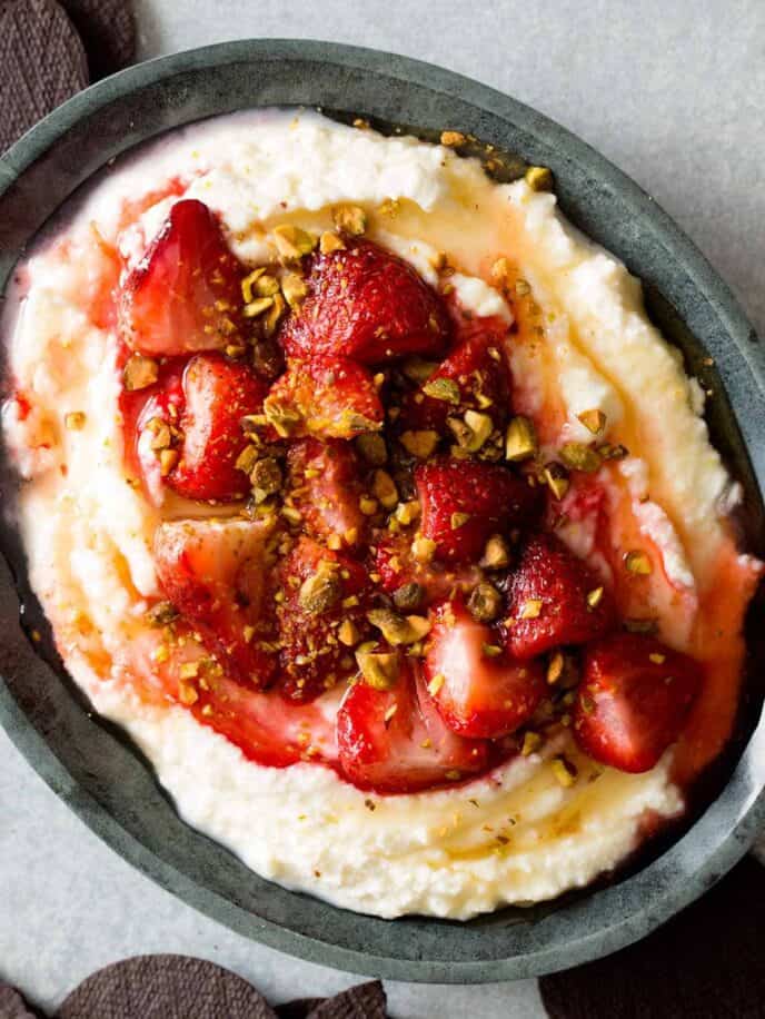 Honey whipped ricotta in a bowl topped with roastes strawberries and pistachios. 