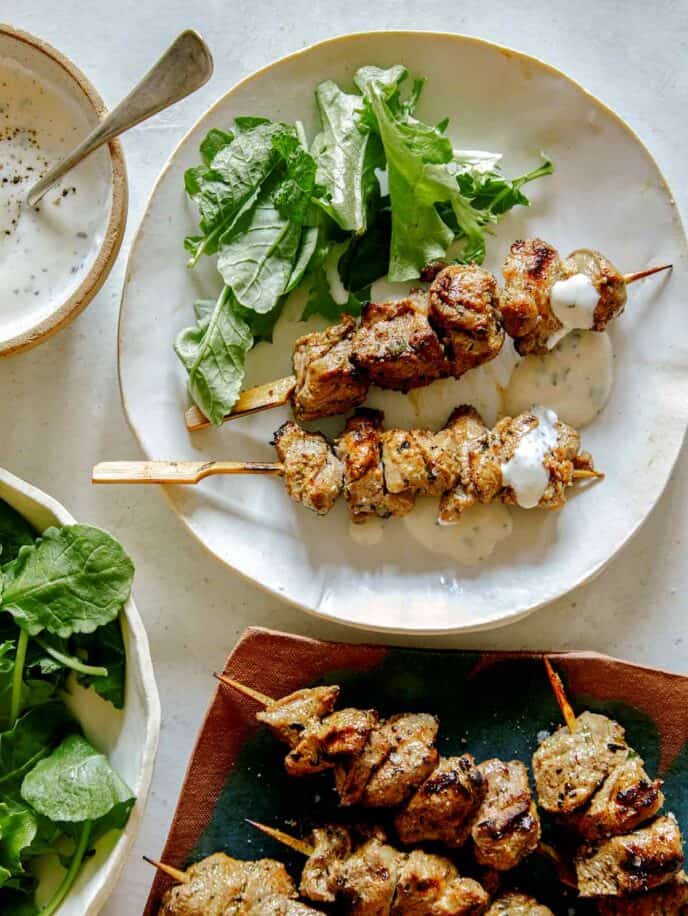 Lamb Kabobs on a plate with drizzled with a yogurt sauce, with a platter of kabobs peeking in. 
