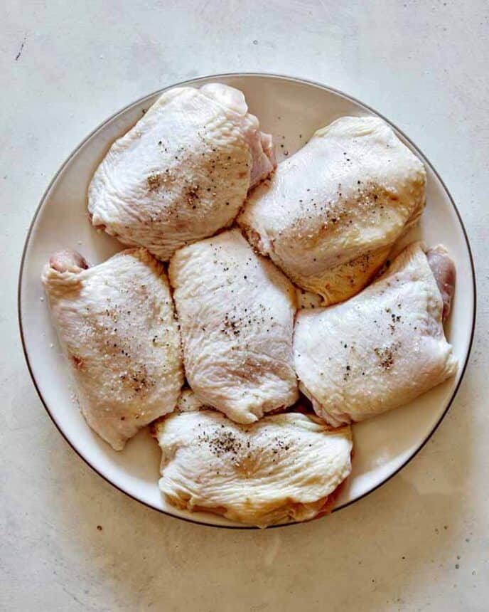 Seasoned raw chicken thighs on a plate with salt and pepper.