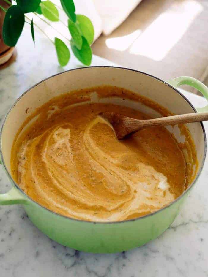 A pot of soup on a counter with cream being stirred in and a wooden spoon.