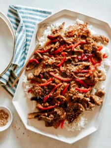 A platter of Szechuan Beef recipe with plates next to it and a ramekin of sesame seeds.