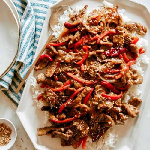 A platter of Szechuan Beef recipe with plates next to it and a ramekin of sesame seeds.