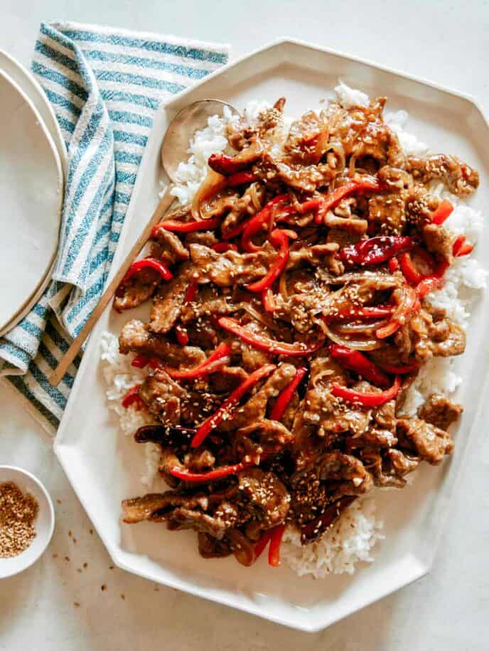 A platter of Szechuan Beef recipe with plates next to it and a ramekin of sesame seeds. 