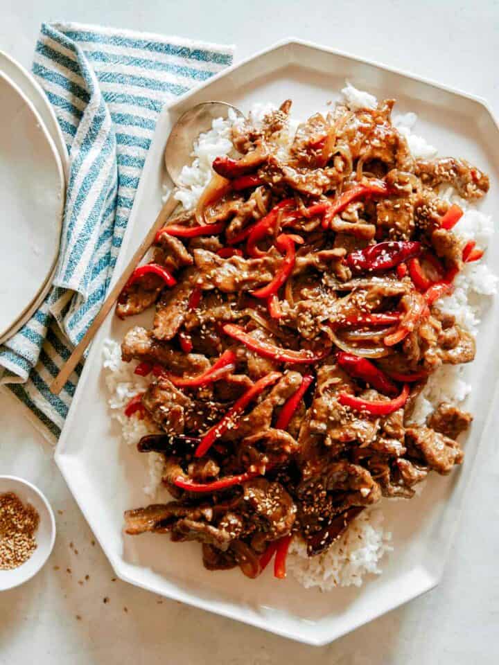 A platter of Szechuan Beef recipe with plates next to it and a ramekin of sesame seeds.