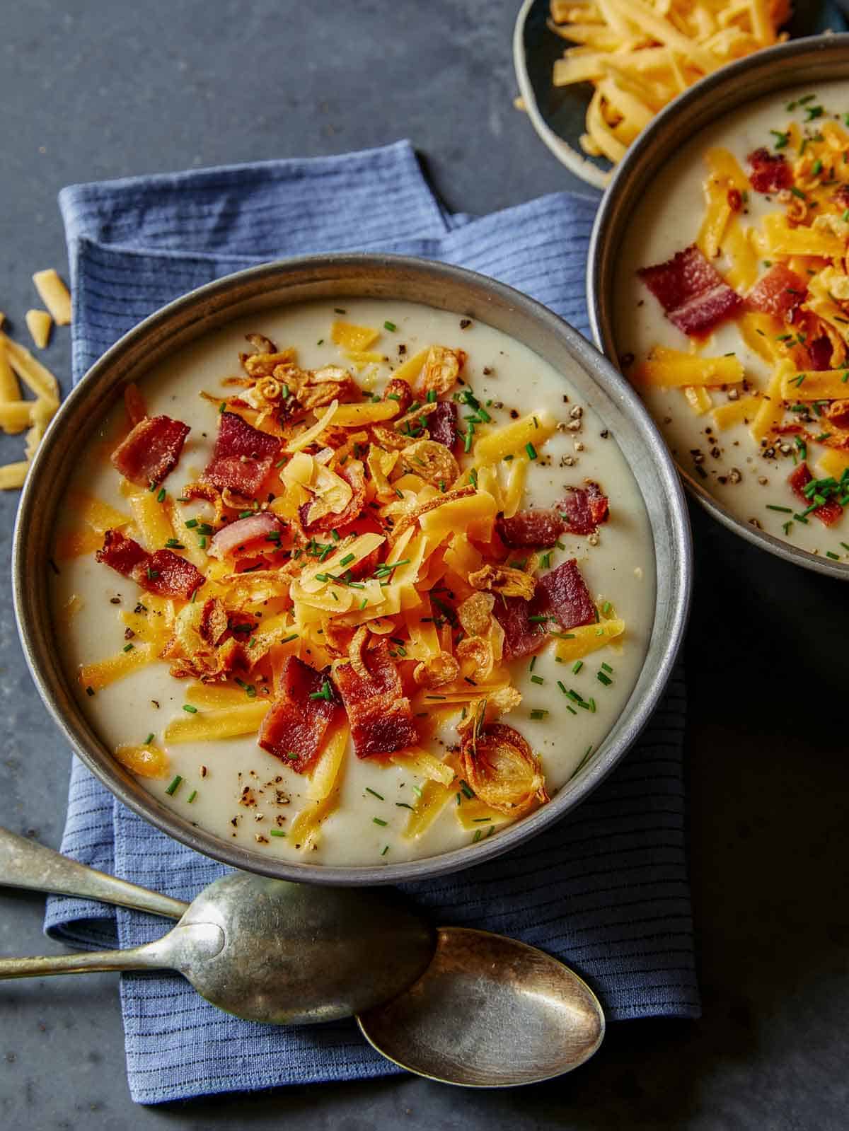 Two bowls of loaded baked potato soup with some shredded cheese next to it.