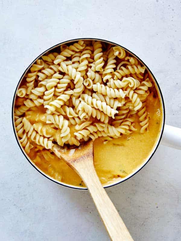 Tossing cooked fusilli pasta into a pot full of pumpkin thyme mac and cheese sauce.