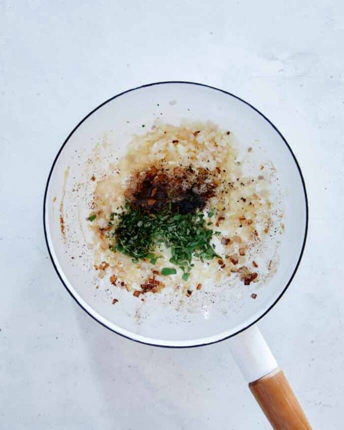 Adding herbs to shallot and garlic in a pot.