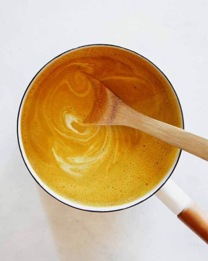 Pumpkin Soup in a stock pot with cream mixed in with a wooden spoon.