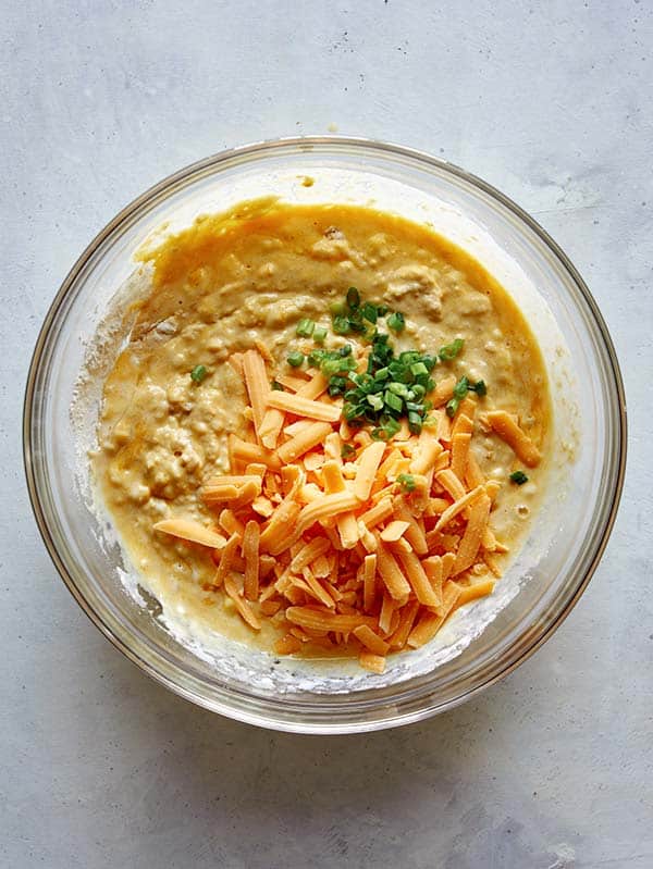 Cheese and green onions added to a pancake batter in a glass bowl.