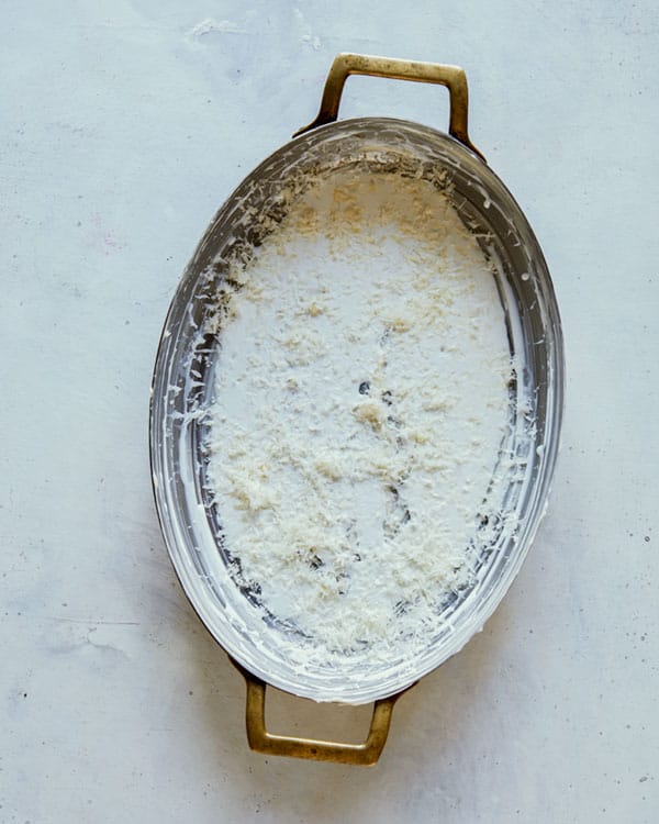 A buttered baking dish with a layer of cream, parmesan, and garlic.
