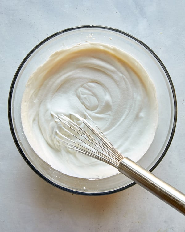 Sweetened whipped cream in a glass bowl with a whisk in it.