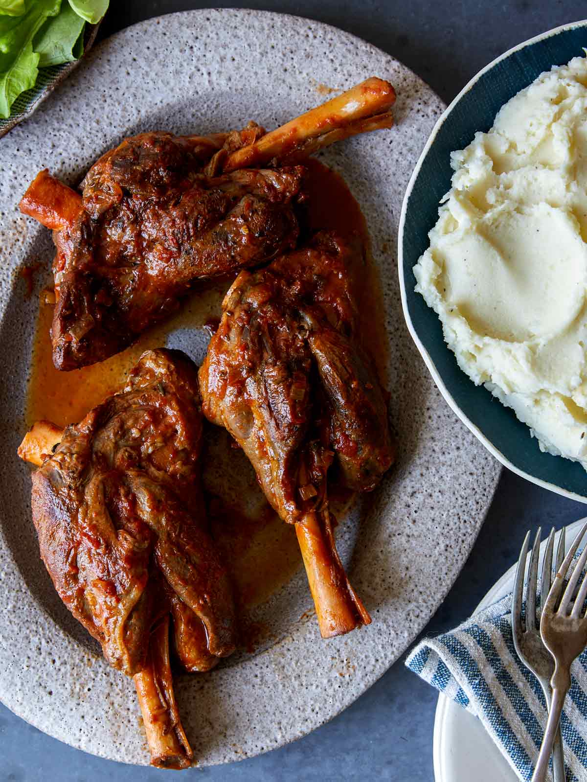 Braised Lamb shanks on a platter with mashed potatoes next to it. 