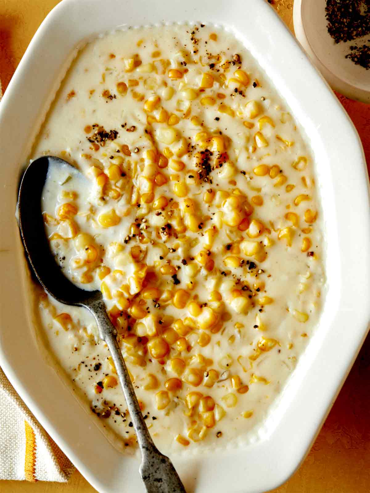 Creamed corn recipe in a serving dish with a spoon in it and fresh pepper on top. 