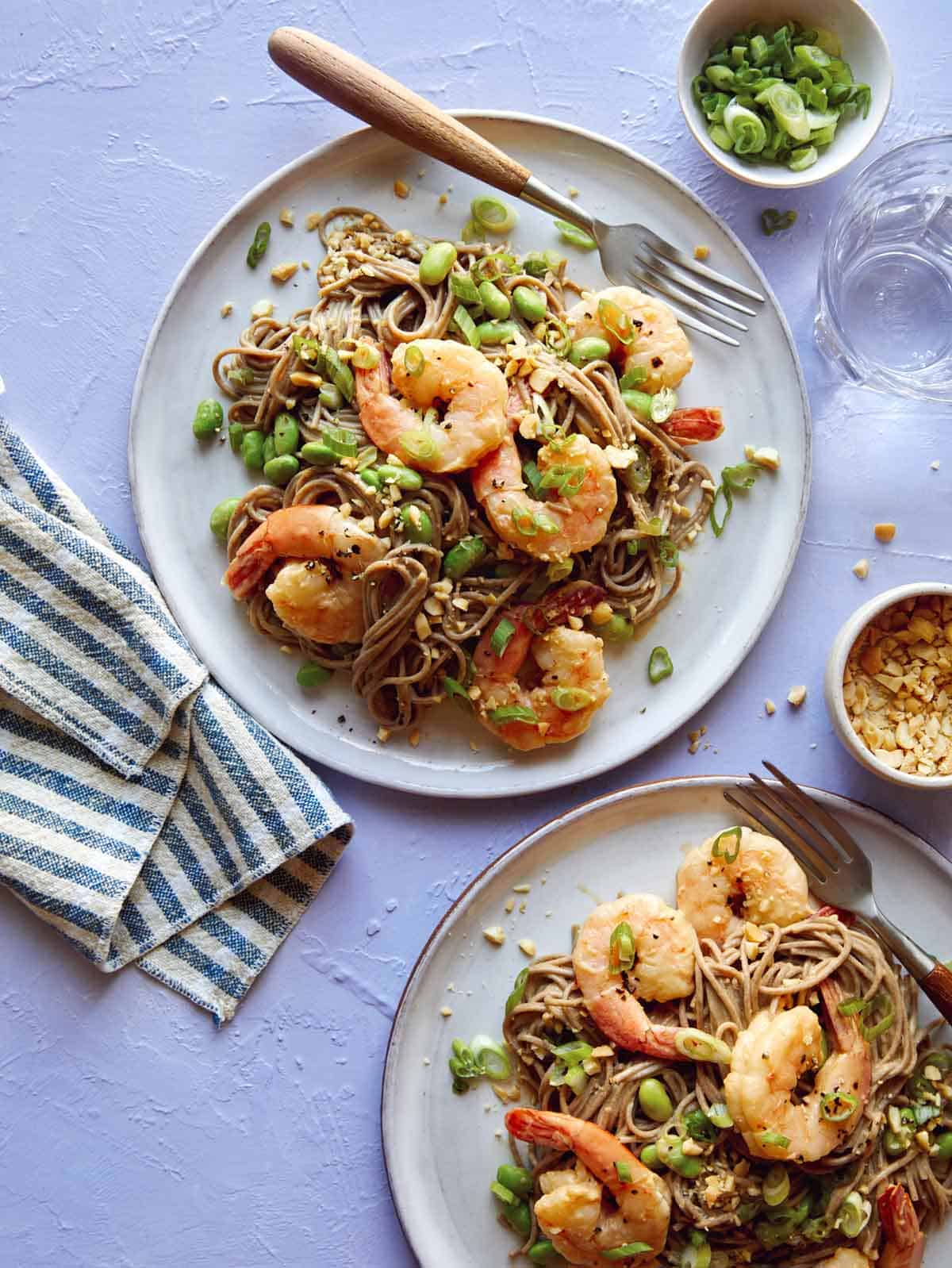 Soba noodles with shrimp on two plates with forks and garnish next to it. 