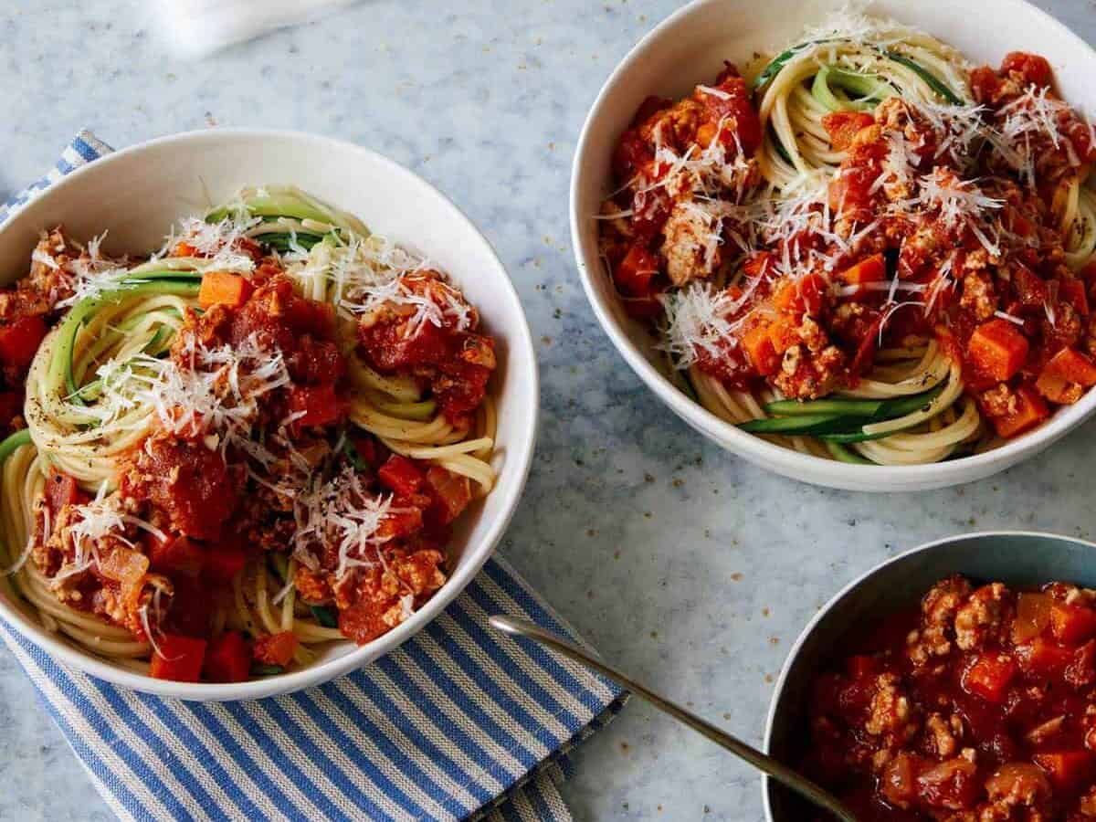 A woman cleaning a spill next to bowls of turkey bolognese with spaghetti and zucchini noodles.