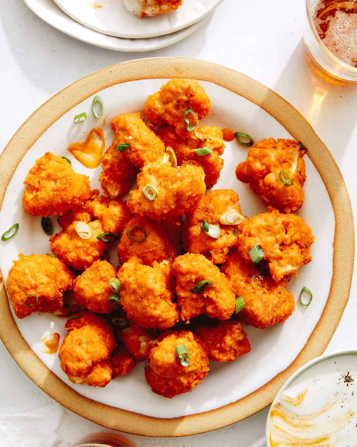 Buffalo cauliflower on a plate with green onions and beer on the side. 