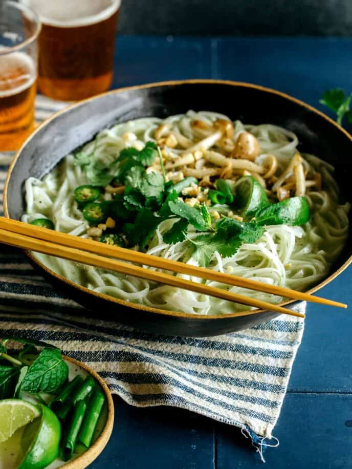 Green curry noodle soup in a bowl with a beer in the background.