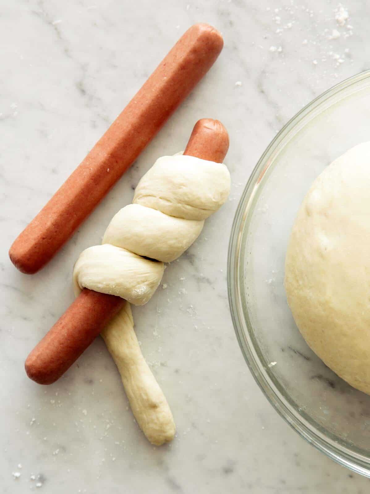 Hot dogs being wrapped in pretzel dough next to a bowl of dough.