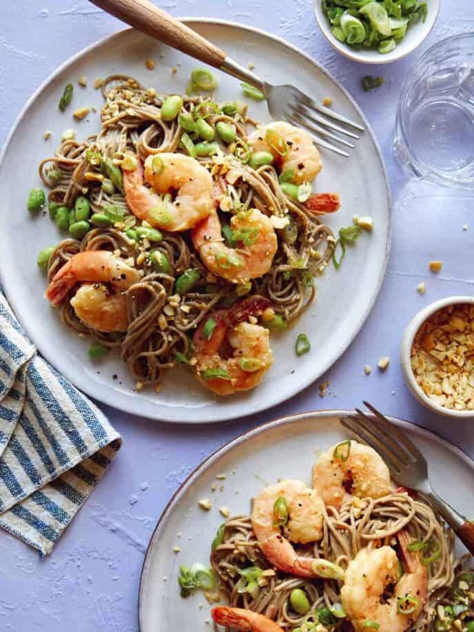 Soba noodles with shrimp on two plates with forks and garnish next to it.