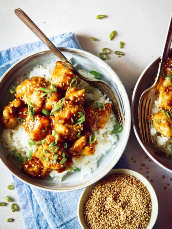 Two bowls of fresh sesame chicken in bowls with rice and a little bowl of sesame seeds.
