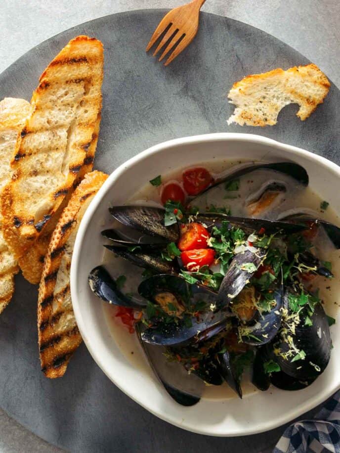 A bowl of white wine steamed mussels on a plate with bread, forks, and drinks.