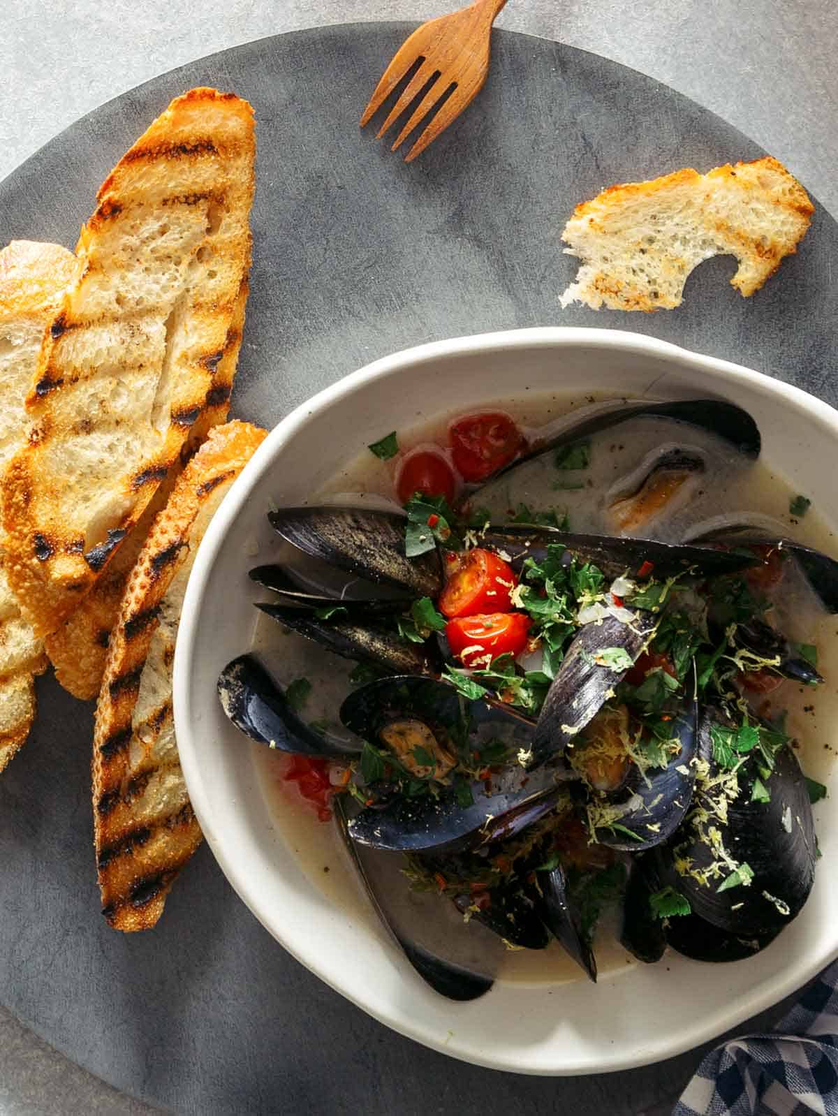 A bowl of white wine steamed mussels on a plate with bread, forks, and drinks.