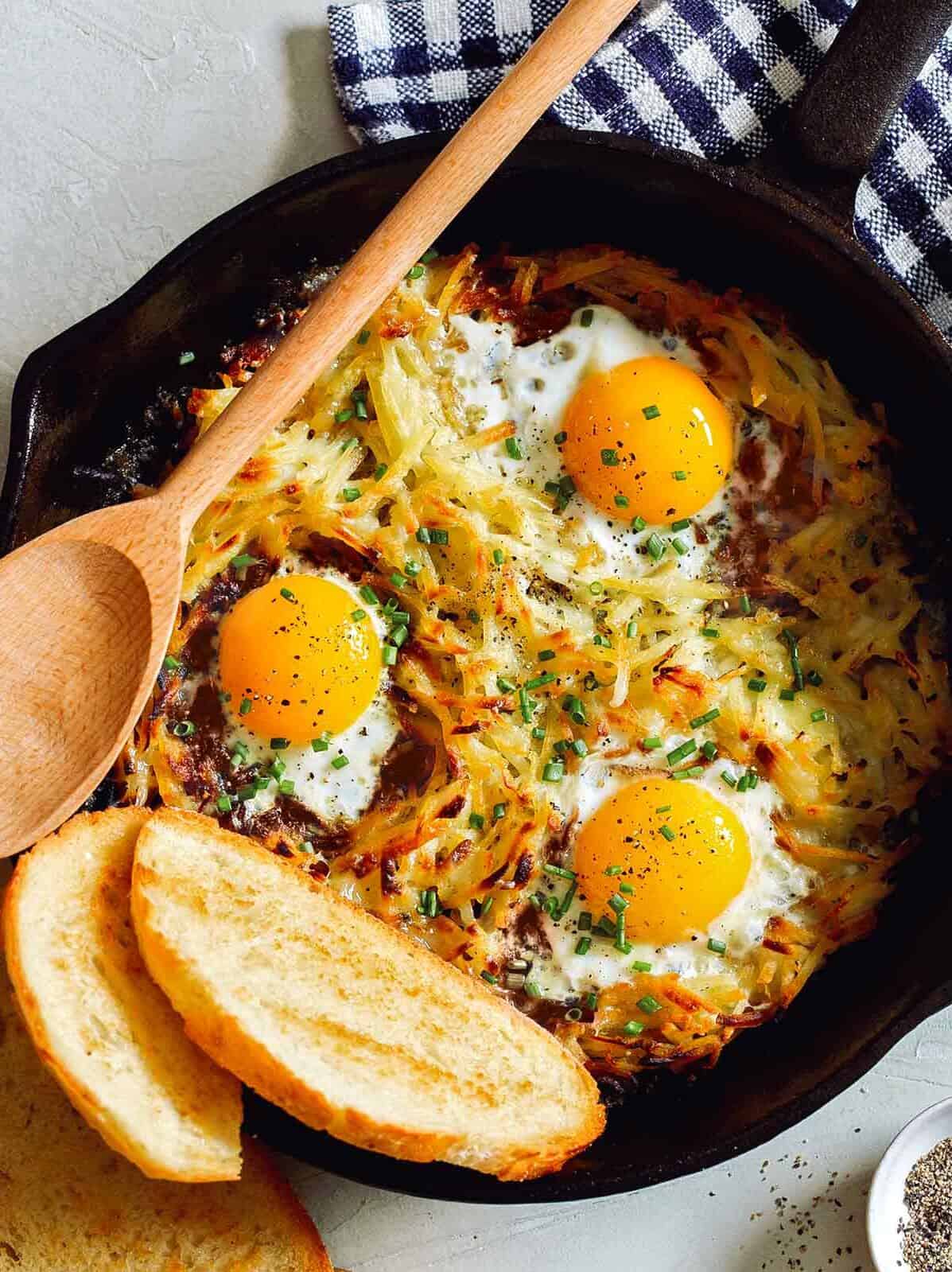 Simple cheesy skillet hash browns and eggs with bread and a wooden spoon.
