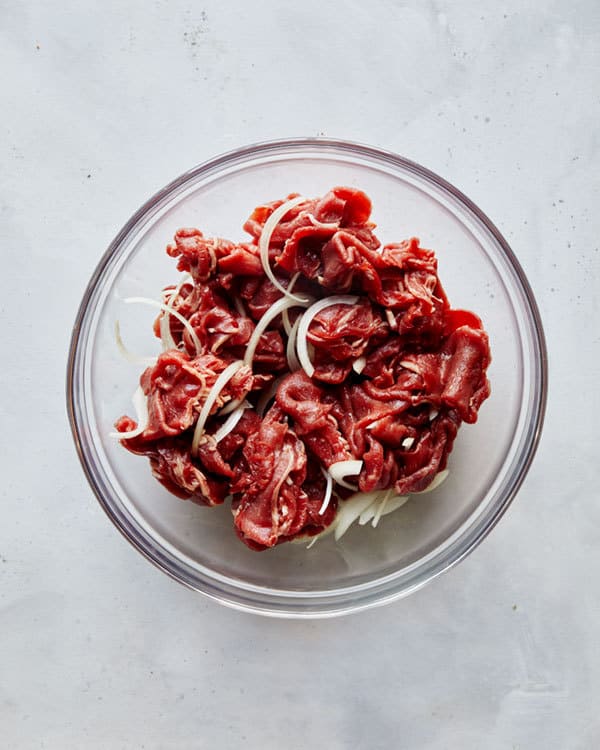 Thinly sliced beef ribeye in a bowl with onion.
