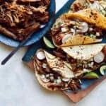 Carnitas recipe in a bowl next to a plate of tacos.