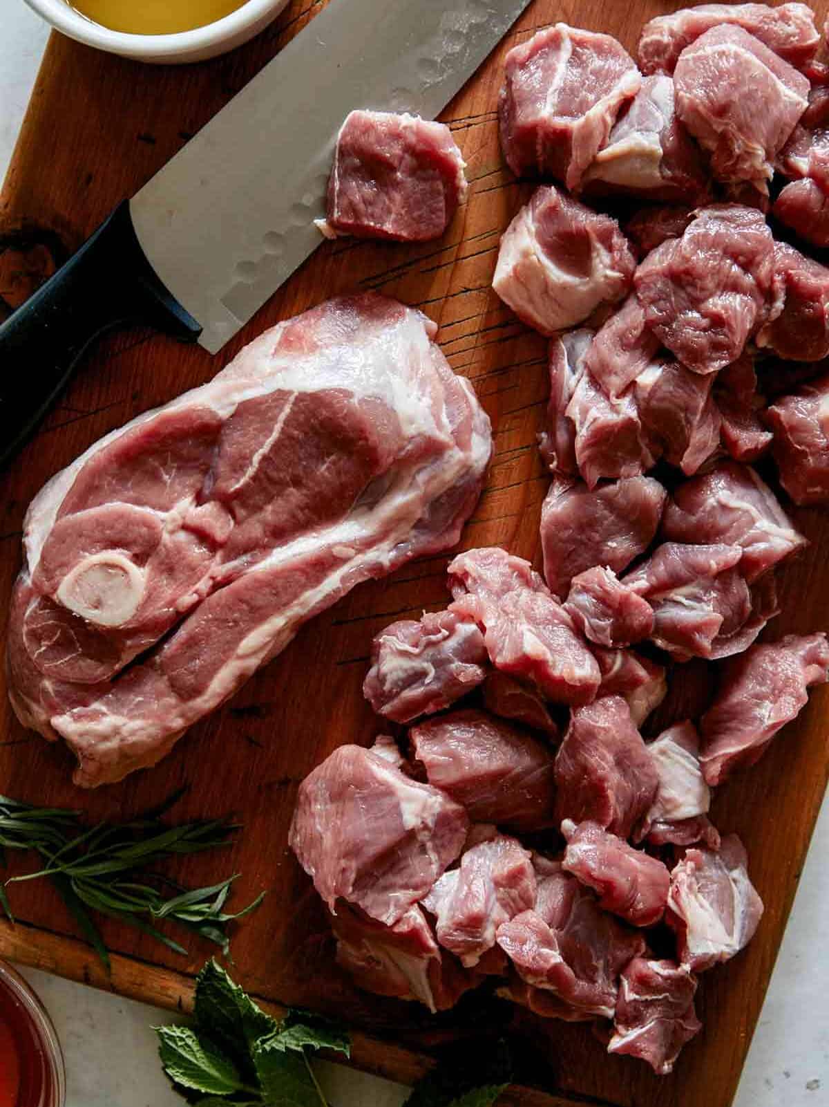 Lamb shoulder cubed on a cutting board with some herbs on the side.