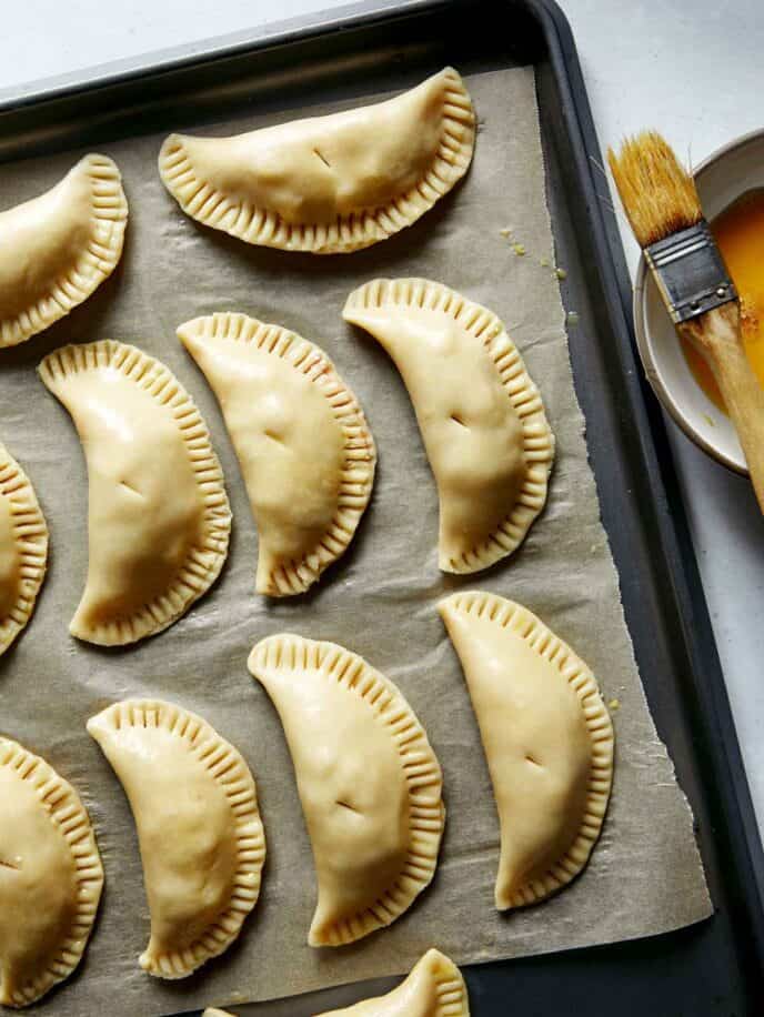 Pork pasties on a baking sheet being brushed with an egg wash.