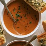 Tomato soup recipe in two bowls with garlic bread on the side.