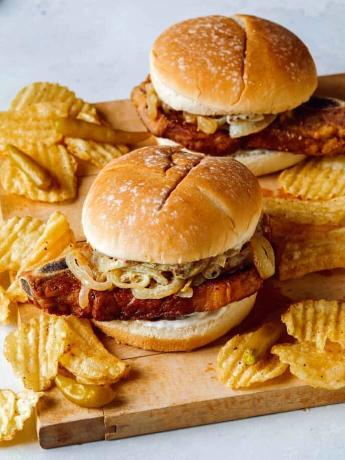 Pork chop sandwiches on a cutting board with potato chips. 