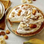 A slice of banana cream pie on a plate with the pie in the background.