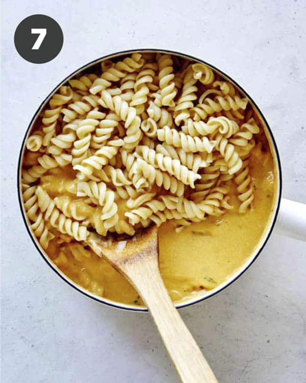 Tossing cooked fusilli pasta into a pot full of pumpkin thyme mac and cheese sauce.