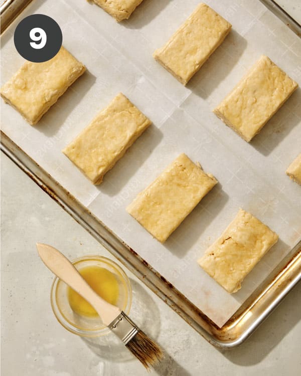 Buttermilk biscuit dough on a baking sheet being brushed with melted butter.