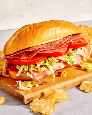 An Italian Grinder sandwich on a cutting board with chips next to it.