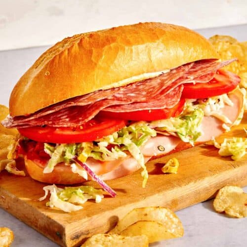 An Italian Grinder sandwich on a cutting board with chips next to it.