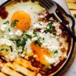 Individual shakshuka with hominy and feta, with a popped egg yolk and grilled flatbread.