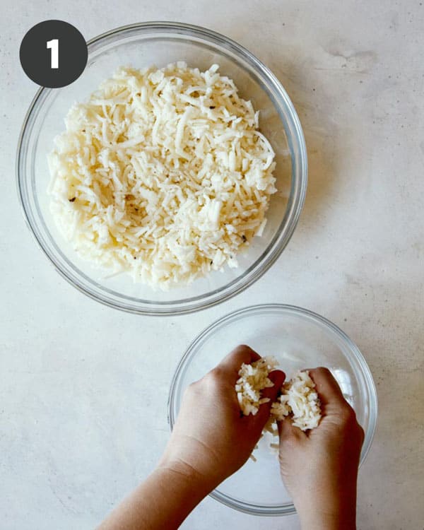 Thawed hash browns in a mixing bowl  with hands squeezing out the moisture. 