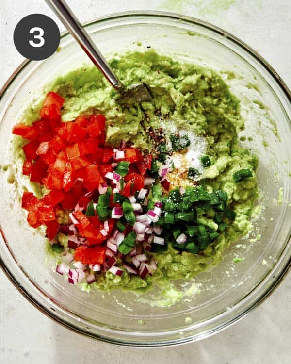 Mashed avocado in a glass bowl with mix ins added. 