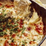A hand with a potato chip dipping into a skillet of crab dip.