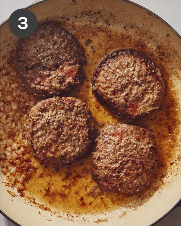 Hamburger patties cooking in a skillet with onions on the side.