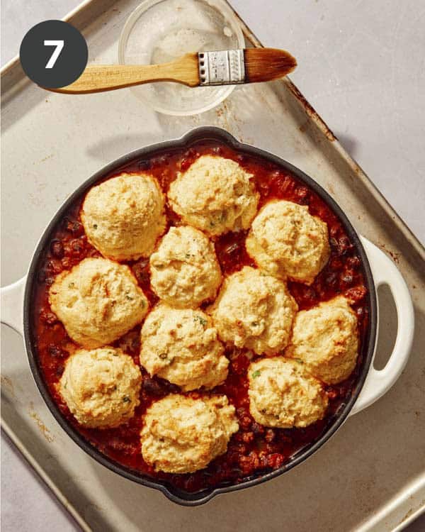 Baked cornbread on top of chili in a skillet with butter being brushed on the cornbread.